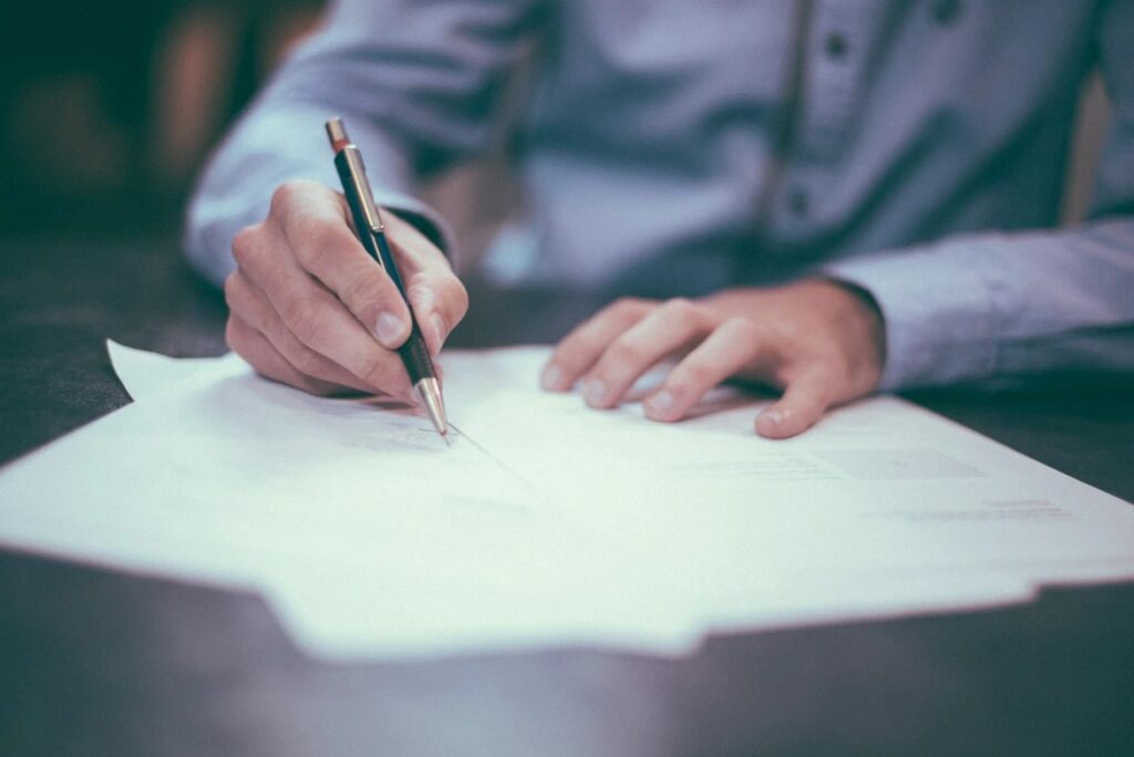  A person is shown signing a document, with a pen in one hand and the other hand holding the document in place. Factors influencing the costs of long term care insurance are displayed on the paper.